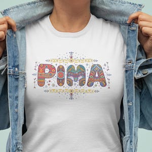 以下が含まれることがあります： カラフルな模様の文字で「PIMA」と書かれた白いTシャツ。文字は赤、オレンジ、青、緑で、上下に装飾的な要素があります。デニムジャケットを着ている人。