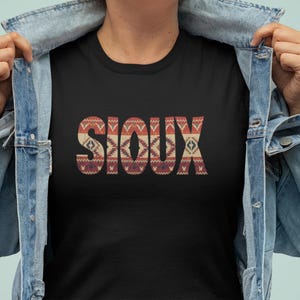 May include: Black t-shirt with the word "SIOUX" in a patterned design. The design incorporates shades of red, beige, and brown. The person is wearing a denim jacket.
