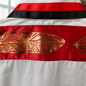 May include: A white shirt featuring a collar with horizontal red and black satin bands. Below the collar, a wide horizontal red satin band displays a repeating metallic gold feather pattern with intricate swirling and dotted line details.