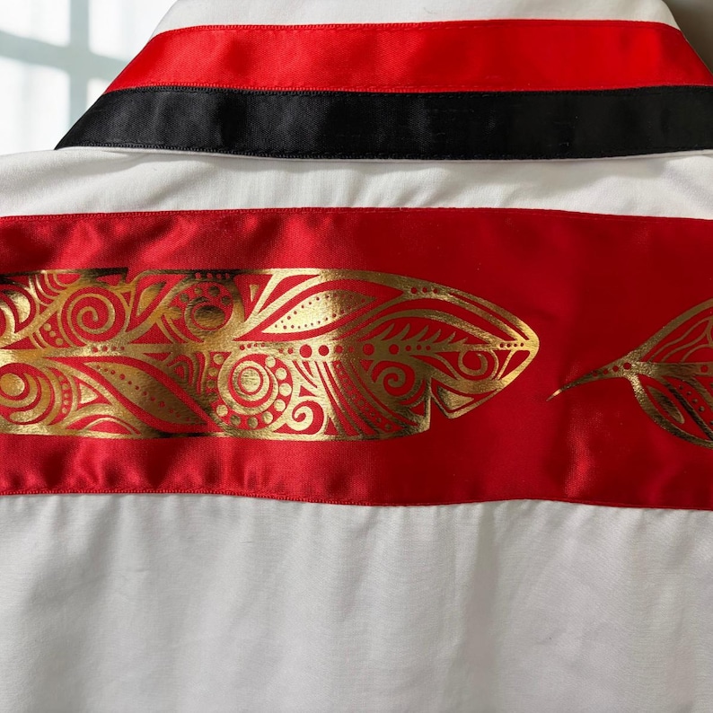 May include: A white shirt featuring a collar with horizontal red and black satin bands. Below the collar, a wide horizontal red satin band displays a repeating metallic gold feather pattern with intricate swirling and dotted line details.