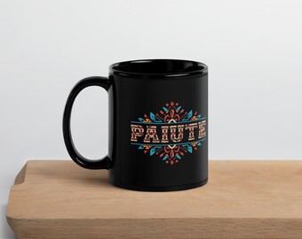 May include: A black ceramic mug with the word "PAIUTE" in a decorative, colorful design. The design includes orange, teal, and red elements. The mug is on a wooden surface.