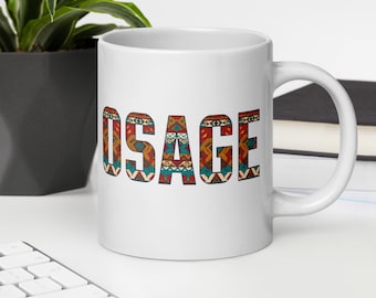 May include: White ceramic mug with the word "OSAGE" in large, colorful letters. The letters are filled with a patterned design in shades of red, orange, teal, and white. The mug has a curved handle and sits on a white surface.