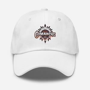 May include: A white baseball cap featuring the word "Comanche" in a stylized font, with a Native American-inspired graphic. The graphic includes a headdress, feathers, and a smiling face. The cap has a curved brim.