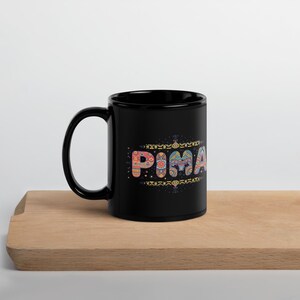May include: A black coffee mug displaying the word "PIMA" in a vibrant, patterned design. The mug has a large handle and is placed on a wooden surface. The design incorporates various colours and geometric shapes.