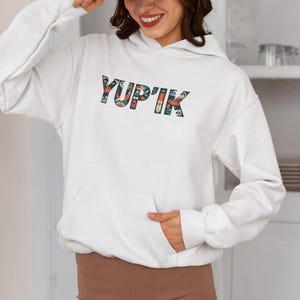 May include: White hoodie with a front pocket and the word "YUP'IK" printed across the chest in a colorful, patterned design. The hoodie has long sleeves and a drawstring hood.