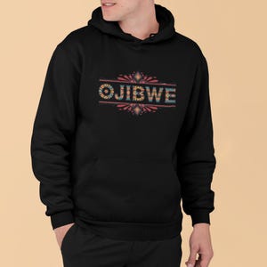 Puede incluir: Sudadera con capucha negra con la palabra "OJIBWE" en letras coloridas y estampadas en el pecho. El diseño presenta un borde decorativo con detalles en rojo, azul y amarillo. La sudadera tiene un bolsillo delantero y una capucha.