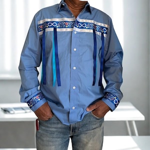 May include: A light blue button-down shirt with decorative ribbon accents. The shirt features white, blue, and teal ribbons, with floral and infinity symbol patterns. The cuffs and upper chest have detailed embroidery. The shirt is paired with blue jeans.