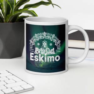 Op de afbeelding: Witte keramische mok met de tekst "Proud Eskimo" in het wit, tegen een donkergroene en blauwe achtergrond met een decoratief ontwerp. De mok staat op een wit oppervlak.