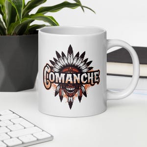 May include: White ceramic mug featuring a graphic of a Native American headdress and the word "Comanche" in orange and black. The design includes feathers and a stylized face. The mug has a curved handle.