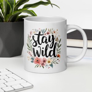 Puede incluir: Taza de cerámica blanca con las palabras "Stay Wild" en escritura negra, rodeada por una corona de flores y vegetación de acuarela. La taza tiene un asa curva y está sobre una superficie blanca.