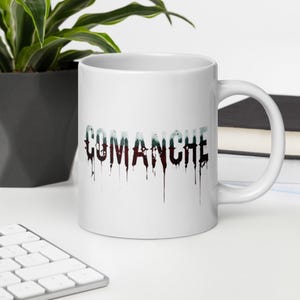 May include: White ceramic mug with the word "COMANCHE" in a dripping, stylized font. The text is a mix of dark red, black, and teal colors. The mug has a standard handle and is set against a white background.