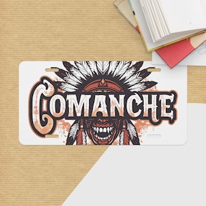 May include: White license plate with the word "COMANCHE" in a distressed font. The design includes a Native American headdress and a stylized face with sharp teeth. The color palette features shades of brown, orange, and white.