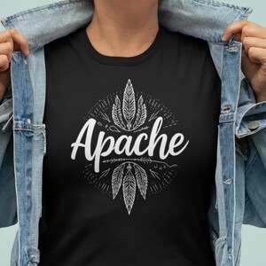 Puede incluir: Camiseta negra con la palabra "Apache" en escritura blanca, rodeada por un diseño circular de plumas y flechas. La persona lleva una chaqueta vaquera.
