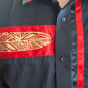 May include: A black button-down shirt featuring a horizontal red satin band across the chest adorned with a gold metallic feather-shaped print. The shirt includes a vertical red satin stripe along the button placket and a red accent on the collar.