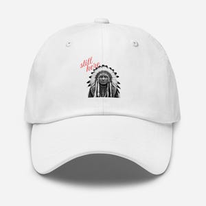 May include: White baseball cap with a black and white illustration of a Native American chief wearing a headdress. The red cursive text "still here" is on the left side. The cap has a curved peak and a classic six-panel design, suitable for various head sizes.
