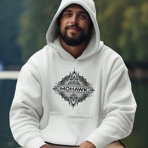Peut inclure: Un sweat à capuche blanc avec le mot "MOHAWK" dans un motif circulaire sur le devant. Le motif est noir et complexe. La personne porte un bracelet et un pantalon kaki.