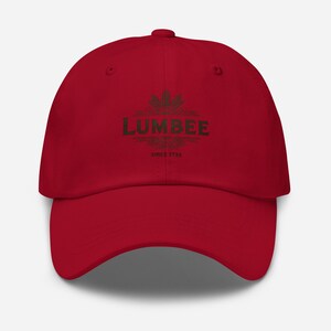 以下が含まれることがあります： 赤い野球帽。装飾的なフォントで「LUMBEE」の文字、その下に「SINCE 1725」と書かれています。帽子はカーブしたつばと調節可能な留め具が付いています。