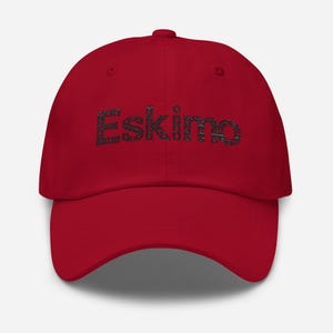 May include: A red baseball cap with the word "Eskimo" embroidered in black, stylized lettering across the front. The cap has a curved brim and a fabric strap closure at the back. This hat is a casual accessory.