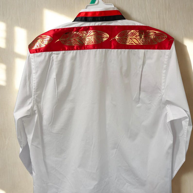 May include: A white long-sleeve button-down shirt featuring a horizontal red band across the upper back and shoulders. The red band is decorated with a repeating gold metallic feather pattern. The collar includes a black and red striped trim.