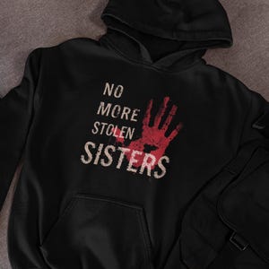 May include: Black hoodie featuring the text "NO MORE STOLEN SISTERS" in a distressed white font. A red handprint graphic is above the word "SISTERS". The sweatshirt has a front pocket and a hood.