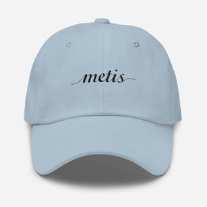 May include: A pale blue baseball cap featuring the word "metis" in black cursive script. The cap has a curved peak and a classic six-panel construction.