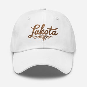 Puede incluir: Gorra de béisbol blanca con la palabra "Lakota" en escritura marrón en la parte delantera. El texto está acompañado de un diseño floral decorativo. La gorra tiene una visera curva y una correa ajustable.