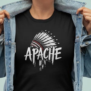 Puede incluir: Camiseta negra con un diseño de tocado nativo americano blanco y rojo y la palabra "APACHE" en una fuente desgastada. La camiseta se lleva debajo de una chaqueta vaquera azul claro.