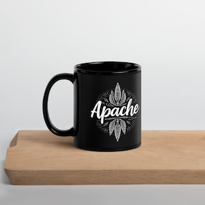 May include: A black ceramic mug with a white design featuring the word "Apache" in a script font, surrounded by stylized feathers. The mug is on a wooden surface.