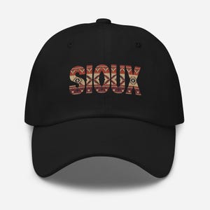 Puede incluir: Gorra de béisbol negra con la palabra "SIOUX" en un diseño estampado. El diseño utiliza tonos de rojo, marrón y crema, creando una apariencia texturizada. La gorra tiene una visera curva y una correa ajustable.