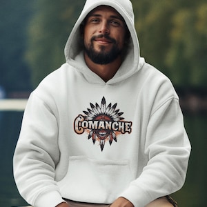 May include: White hooded sweatshirt with the word "COMANCHE" in an orange and brown font, above a graphic of a Native American headdress. The sweatshirt has a front pocket and is being worn by a person.