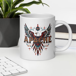 May include: White ceramic mug with a detailed eagle graphic, the word "APACHE", and decorative elements in red, teal, and brown. The mug has a curved handle and is suitable for hot or cold drinks. The design is reminiscent of Native American art.
