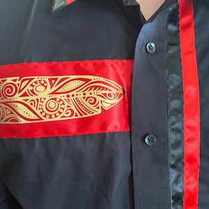 May include: A black button-down shirt featuring a horizontal red satin band across the chest with a gold-colored feather graphic. The shirt includes a vertical red satin stripe along the button placket and black buttons down the front.