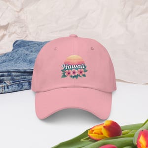 May include: A light pink baseball cap featuring a graphic of a sunset over the ocean with the text Hawaii written in white script above three pink hibiscus flowers and green leaves centered on the front panel of the six-panel crown.