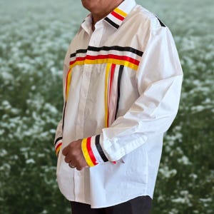 May include: A white long-sleeve button-down shirt featuring horizontal ribbon trim across the chest and collar in black, red, and yellow. Vertical ribbons in the same colors extend downward from the chest trim, and the cuffs feature matching bands.