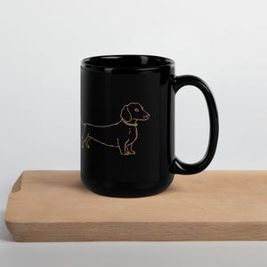 Può includere: Tazza in ceramica nera con un contorno dorato di un cane bassotto. Il cane è raffigurato di profilo con un collare. La tazza è appoggiata su una superficie di legno.