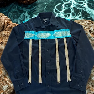 May include: A black button-down shirt with turquoise and gold detailing. The shirt has turquoise fabric across the shoulders and chest, decorated with gold feather patterns. Beige ribbons hang vertically from the chest.