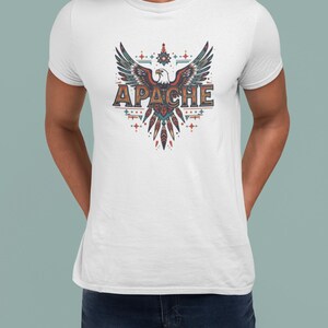 Op de afbeelding: Wit t-shirt met een gedetailleerde afbeelding van een adelaar met uitgespreide vleugels en het woord "APACHE". Het ontwerp bevat tinten rood, blauw en bruin. Decoratieve elementen omringen de adelaar.