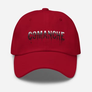 May include: A red baseball cap with the word "COMANCHE" in white and black dripping font across the front. The hat has a curved brim and a fabric strap closure.