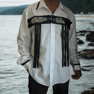 Puede incluir: Camisa blanca abotonada con detalles de plumas y cintas negras. La camisa presenta una banda negra en el pecho con diseños de plumas y cintas negras verticales. El cuello y los puños tienen ribetes negros.