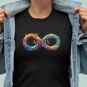 Puede incluir: Camiseta negra con un diseño de símbolo de infinito colorido y la palabra "METIS" en el centro. El símbolo tiene un efecto acuarela con tonos de naranja, amarillo, azul y morado. La persona lleva una chaqueta vaquera.
