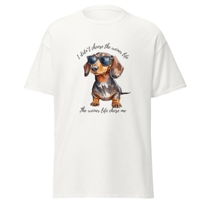 Puede incluir: Camiseta blanca con una ilustración de acuarela de un perro salchicha con gafas de sol. El texto encima del perro dice "No elegí la vida salchicha" y debajo del perro dice "La vida salchicha me eligió".