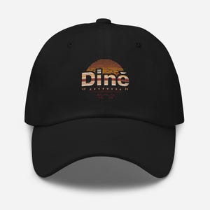 Puede incluir: Gorra de béisbol negra con visera curva. La gorra presenta la palabra "Diné" en una fuente decorativa, sobre un círculo con colores de atardecer. El diseño incluye patrones y símbolos geométricos.