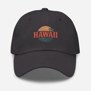 May include: A dark gray baseball cap featuring a distressed graphic of a sunset, palm tree, and ocean waves. The word "HAWAII" is printed in a vintage orange font across the center of the cap. The cap has a curved brim and an adjustable strap.