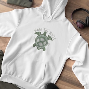 Könnte beinhalten: Weißer Kapuzenpullover mit Kängurutasche, verziert mit einem grünen Schildkröten-Motiv und dem Schriftzug "WHAT THE SHELL". Eine grüne Mütze, schwarze Schuhe und Kopfhörer sind ebenfalls zu sehen.