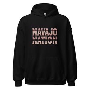 Puede incluir: Sudadera con capucha negra con las palabras "NAVAJO NATION" estampadas en el pecho en un diseño de patrón rosa y blanco. La sudadera tiene un cordón y un bolsillo delantero.