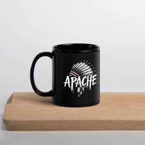 May include: Black ceramic mug featuring a white graphic of a Native American headdress and the word "APACHE". The mug is placed on a wooden surface.