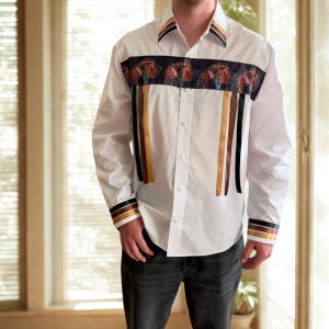 May include: A white, long-sleeved button-down shirt. It has a decorative band across the chest with a repeating saddle pattern. Vertical ribbons in brown, black, and gold are on the front. The collar and cuffs have matching trim.