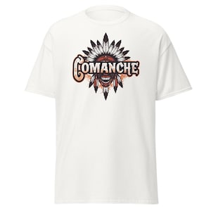 May include: White t-shirt featuring a graphic design with the word "Comanche" in a stylized font. The design includes a Native American headdress and face with a menacing expression, in shades of brown, orange, and black.