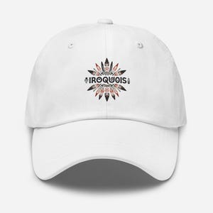 May include: A white baseball cap featuring a circular design with the word "IROQUOIS" in black text. The design includes red, black, and white tribal patterns. The cap has a curved brim and an adjustable strap.