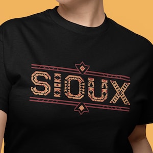 May include: Black t-shirt featuring the word "SIOUX" in a decorative, Native American-inspired design. The text is in a reddish-orange color, with geometric patterns and lines. The shirt is a crew neck.